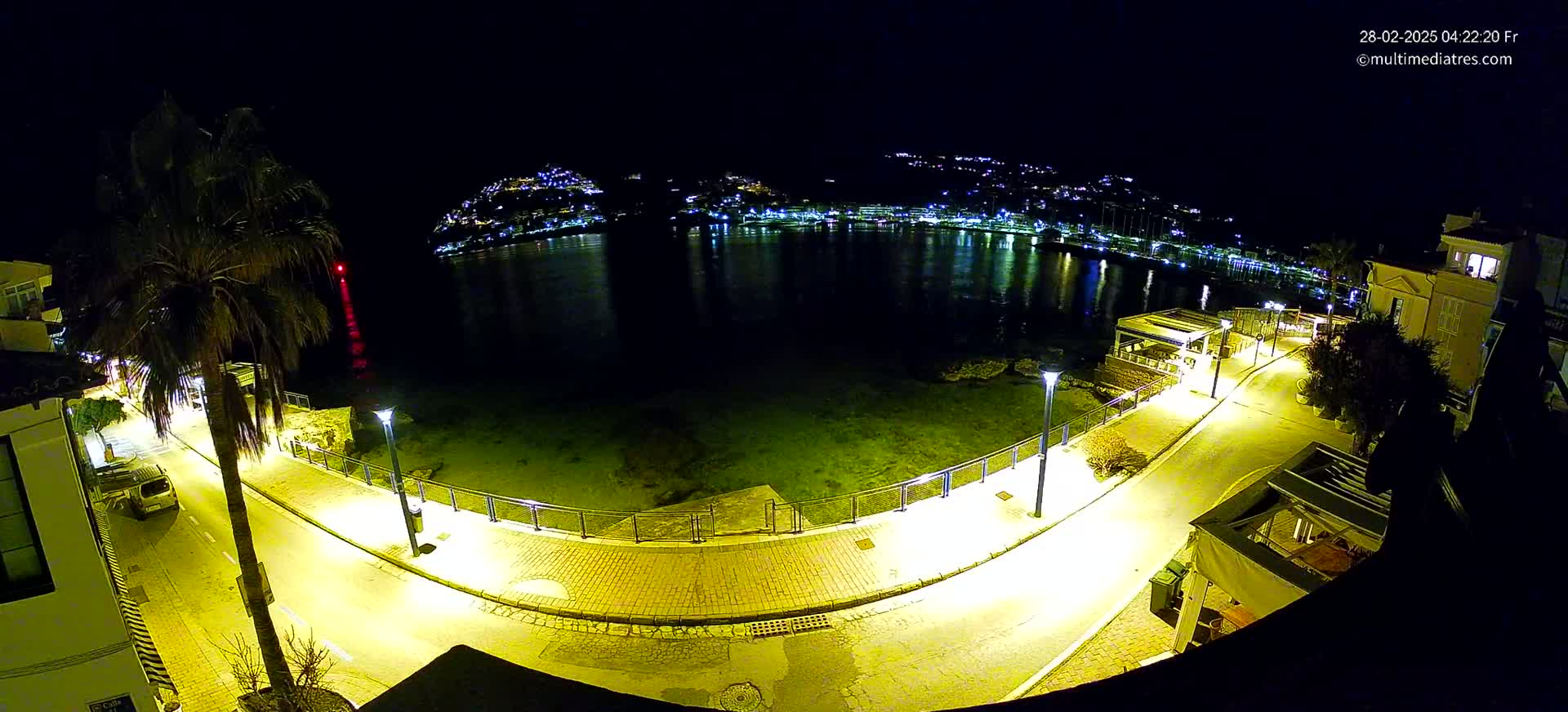 A nighttime, high-angle view of a waterfront promenade illuminated by streetlights, with a calm body of water reflecting the lights of a town on the opposite shore.