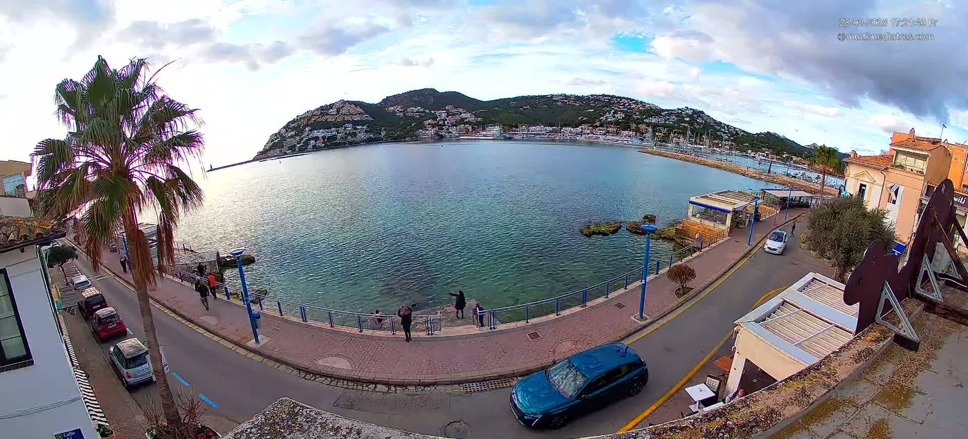 A partly cloudy day overlooks a calm bay with a hilltop town in the background, a curved road alongside the water's edge, and several people standing near a waterfront walkway.