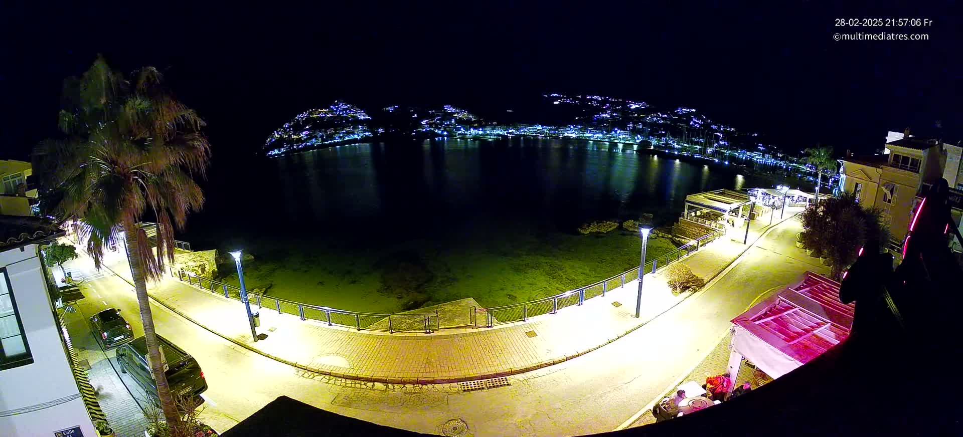 A nighttime, high-angle view shows a calm body of water reflecting the lights of a town on the opposite shore, with a paved walkway and streetlights illuminating a waterfront area featuring palm trees and buildings.