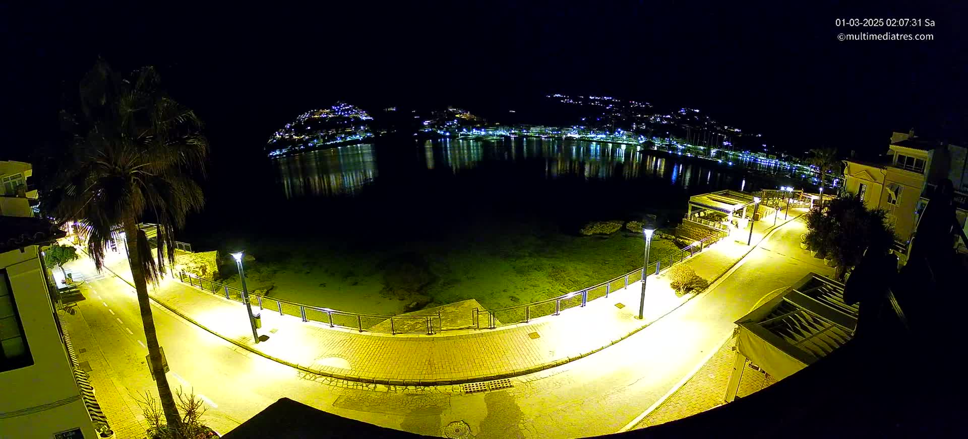 A nighttime, high-angle view shows a brightly lit waterfront promenade curving around a calm bay, with a hilltop town twinkling with lights across the water.