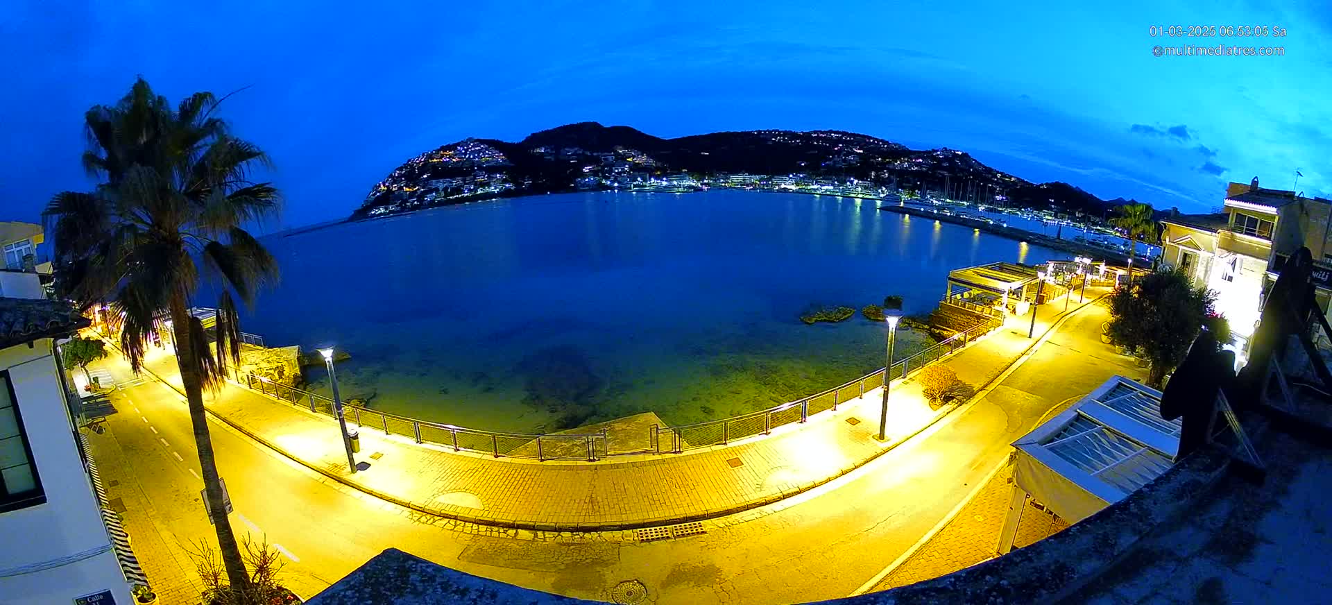 A nighttime, waterfront view shows a curving, illuminated road next to a calm, dark-blue sea, with a hilltop town visible in the distance under a twilight sky.