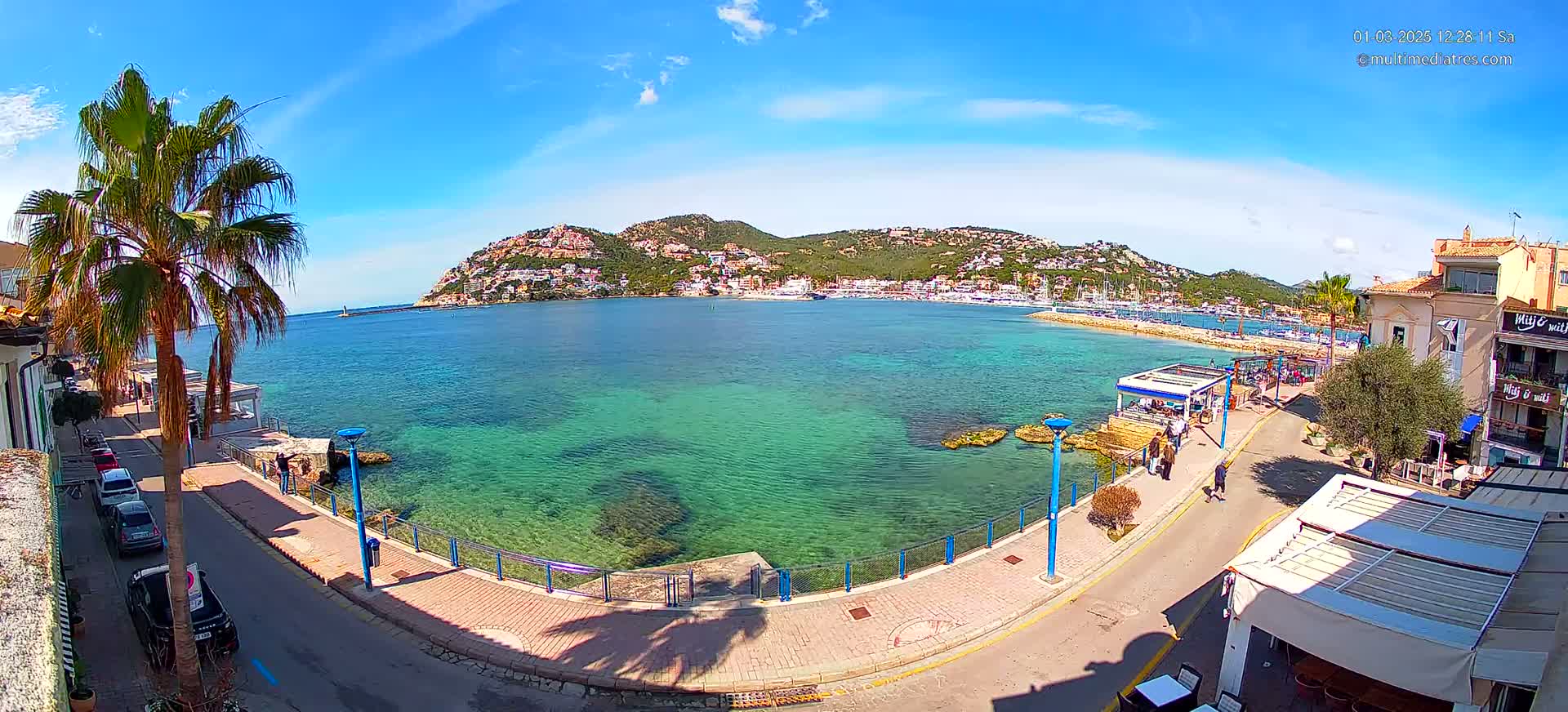 A clear sunny day reveals a picturesque coastal town, with a calm, shallow turquoise bay bordered by a paved walkway and buildings, and a hill dotted with houses in the background.