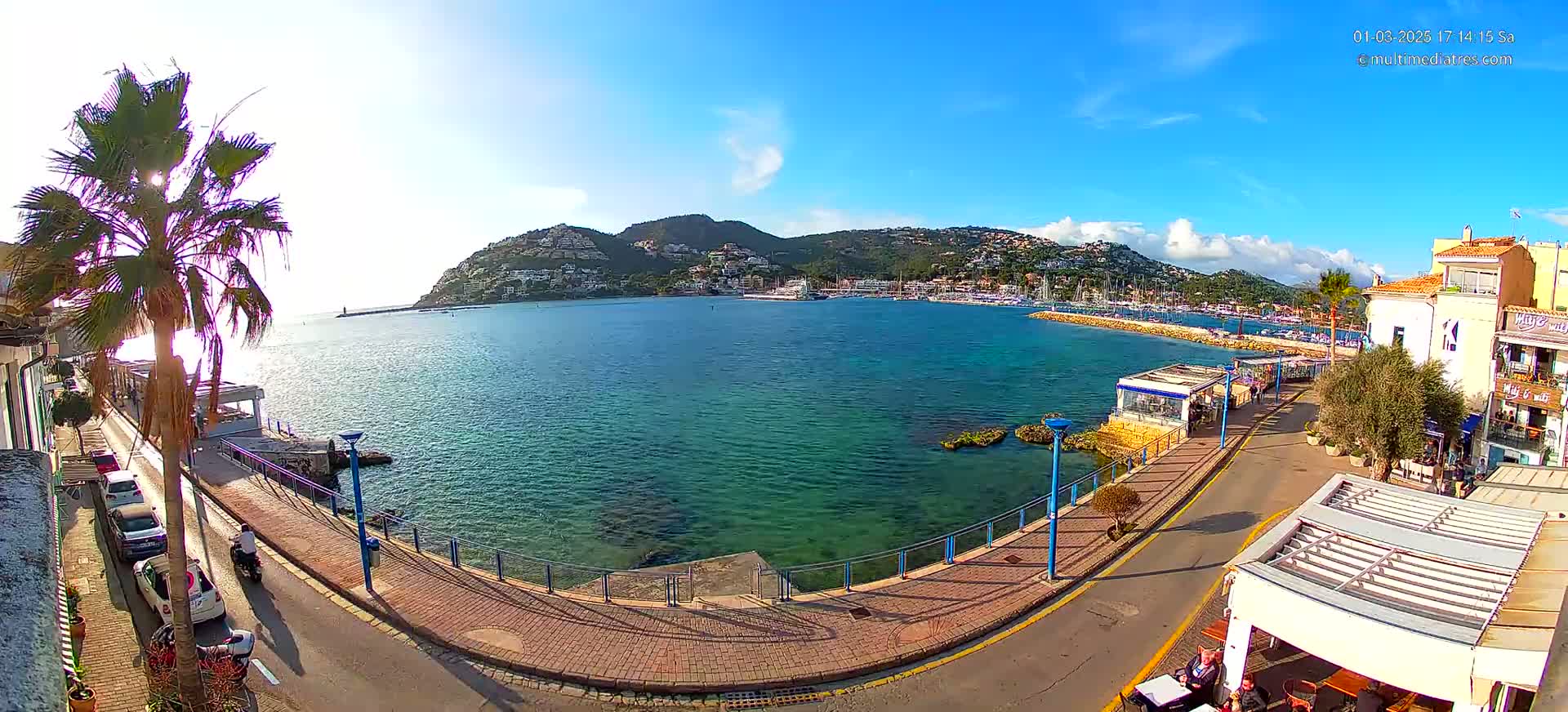 A sunny day overlooking a calm, turquoise bay with a town and hillside buildings in the background, and a palm tree in the foreground.