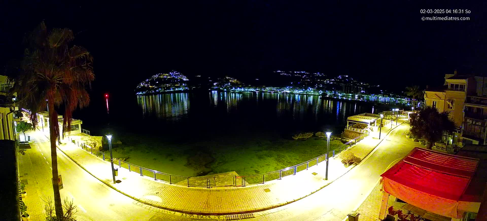 A nighttime view of a waterfront promenade, illuminated by streetlights, with a calm body of water reflecting the lights of a town nestled on a hillside in the distance.