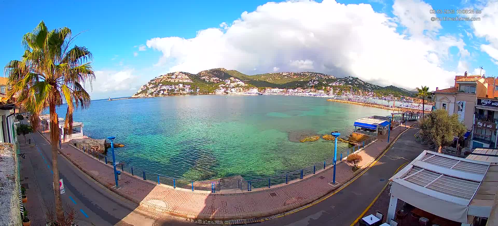 A partly cloudy sunny day reveals a clear, teal bay with a town and hillside of buildings nestled against a mountainous backdrop.