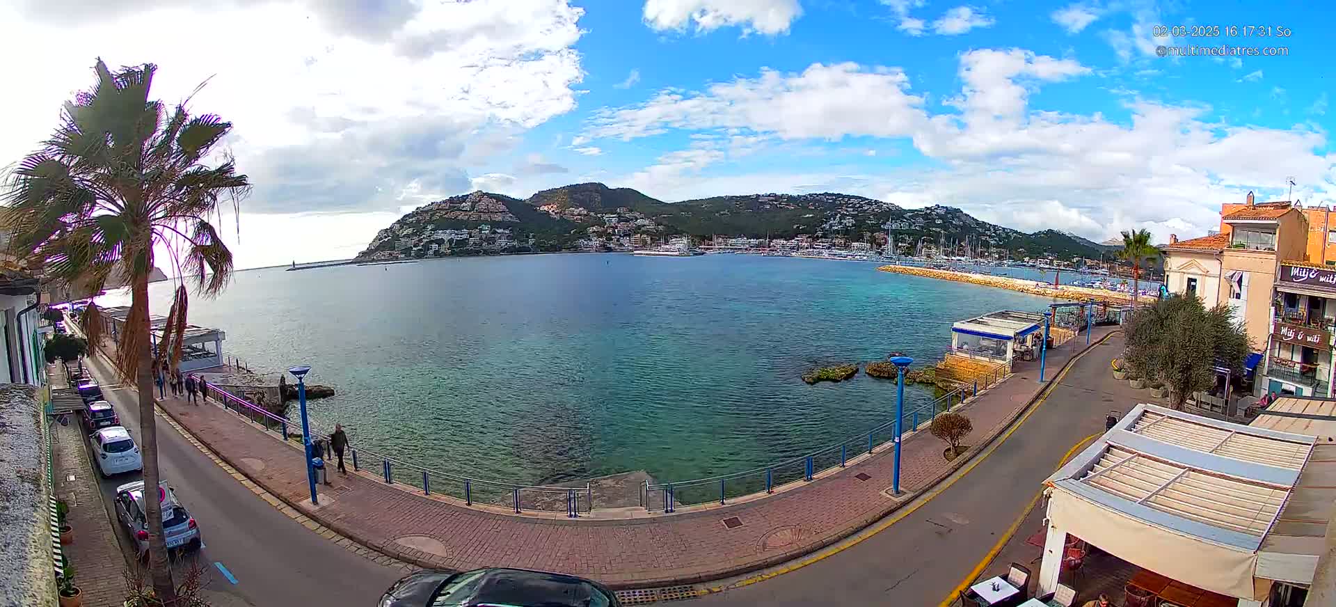 A partly cloudy day shows a harbor filled with boats, with a hill covered in buildings in the background, seen from a seaside promenade with parked cars and people walking.