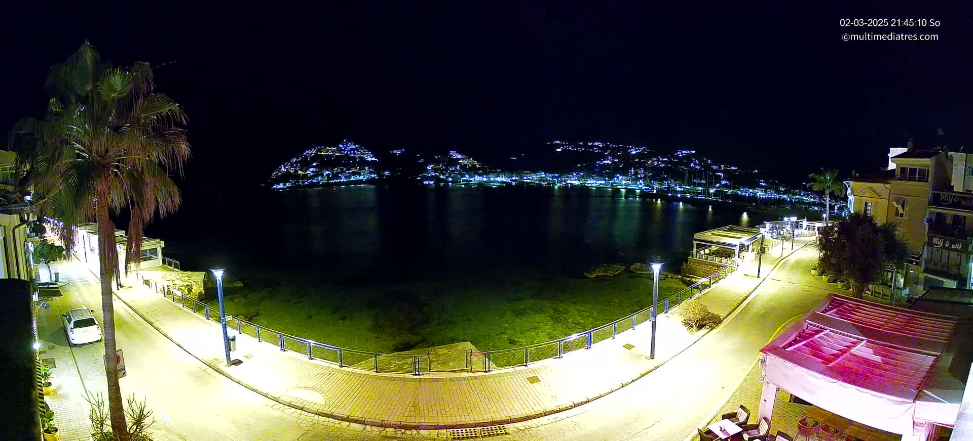 A nighttime view of a waterfront promenade with palm trees, buildings, and a calm bay reflecting the lights of a town across the water.
