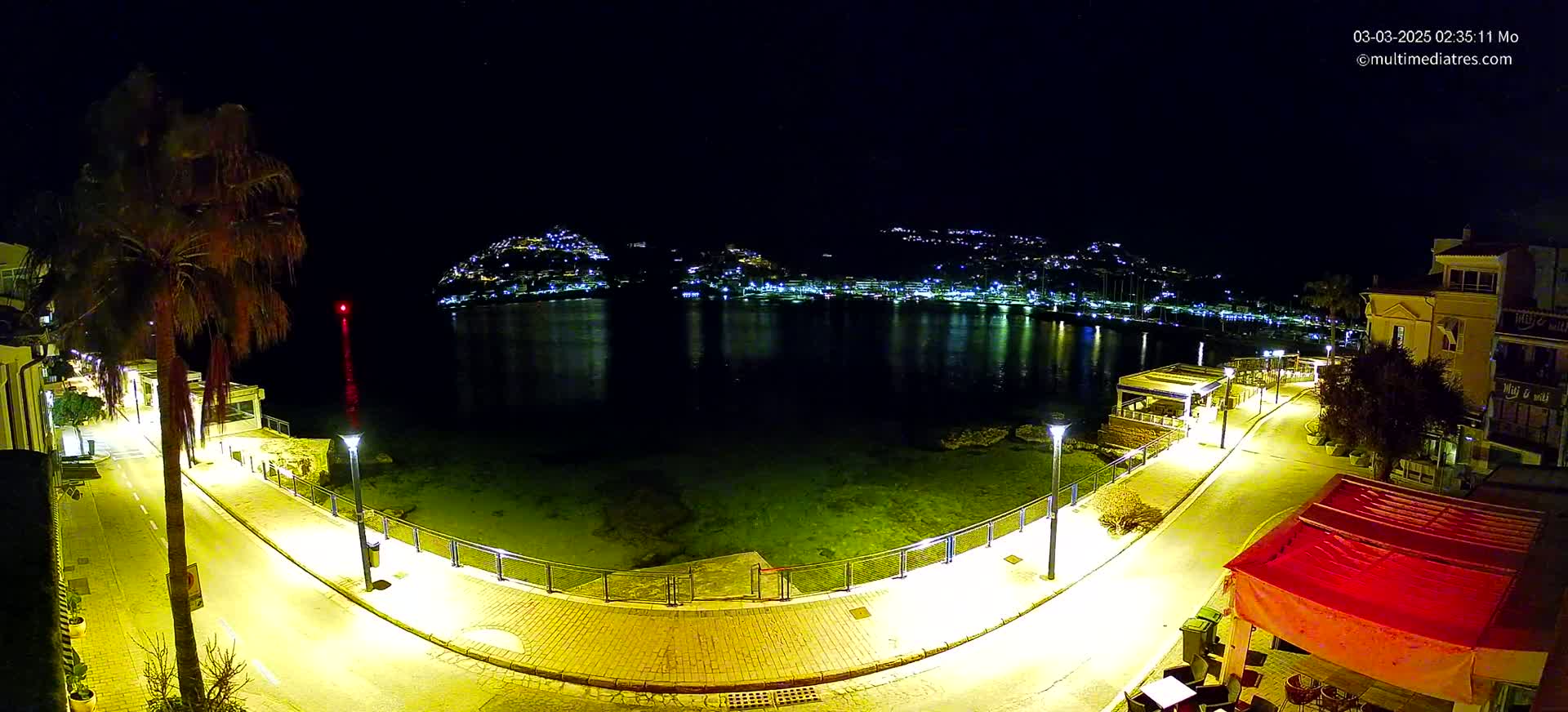 A nighttime, illuminated waterfront path curves around a calm bay, with a hilltop town glowing with lights across the water.