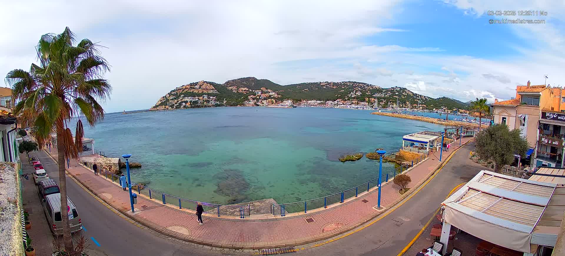 A partly cloudy day shows a coastal town with a calm, clear, teal bay, a paved waterfront walkway, parked cars, and buildings, all nestled beneath a hill covered in houses and greenery.