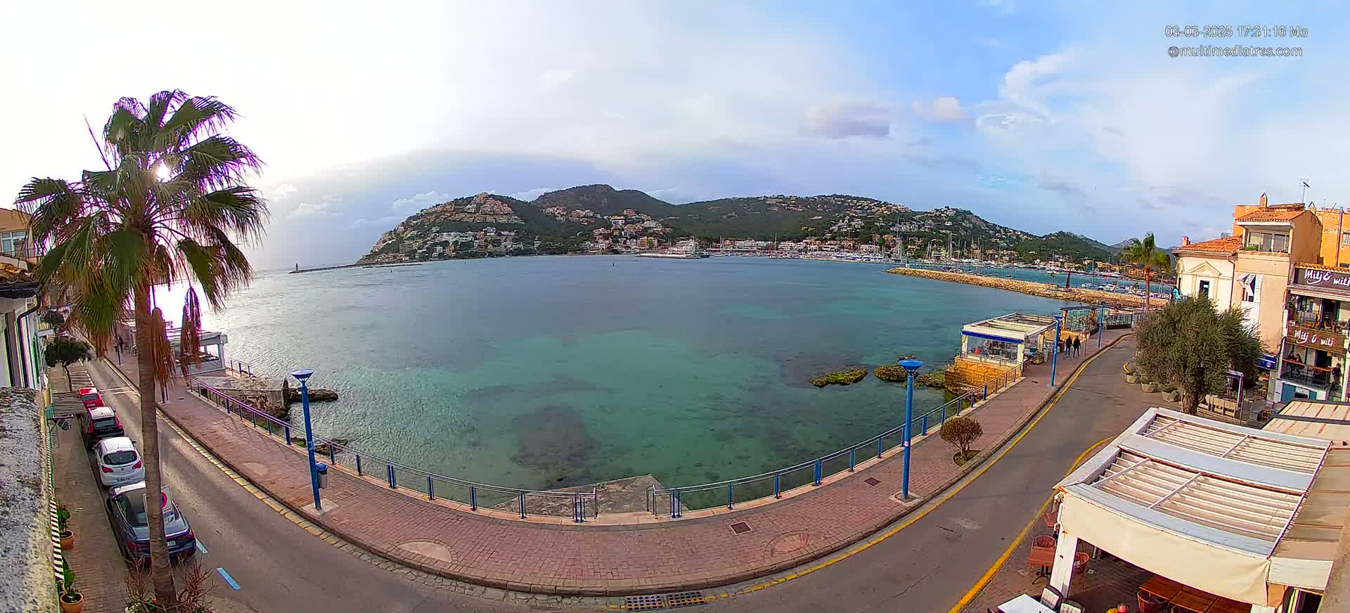 A partly cloudy day reveals a seaside town with a calm, clear turquoise bay, hills covered in buildings in the background, and a palm tree near a paved walkway lined with parked cars and a few buildings.