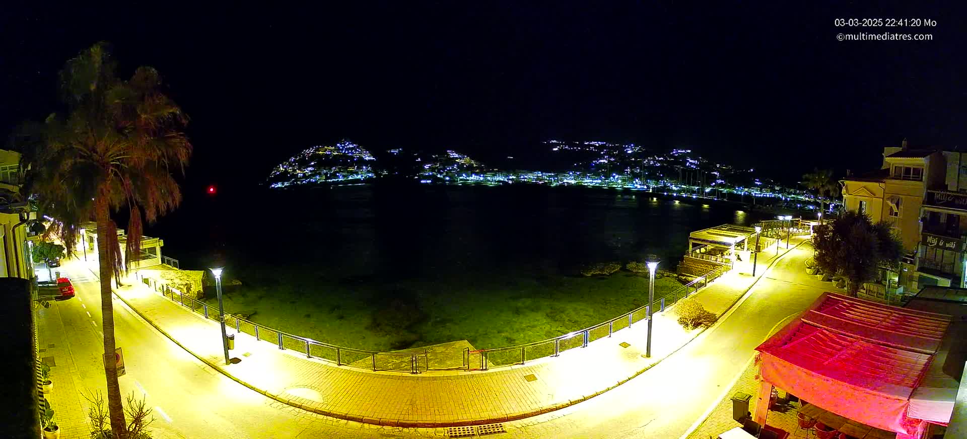 A nighttime view of a waterfront promenade illuminated by streetlights, overlooking a calm body of water and a hilltop town speckled with lights.