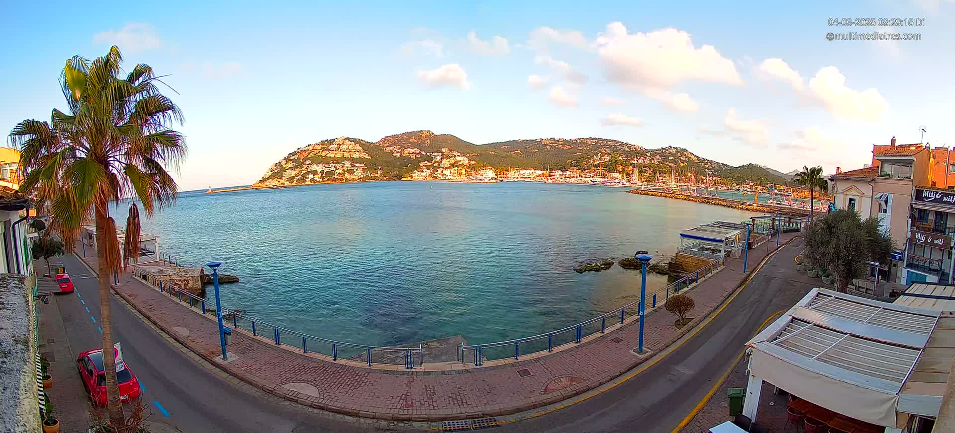 A partly cloudy, sunny day overlooking a calm bay with a town nestled against a hillside, palm trees lining a paved waterfront road.