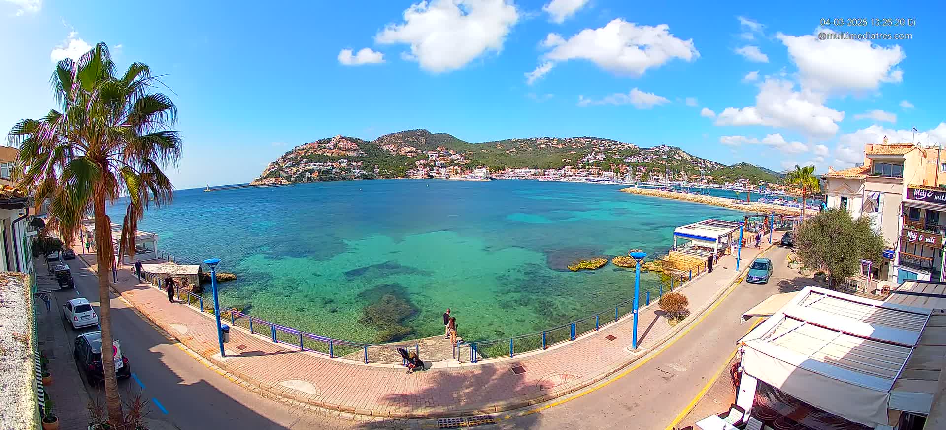 A sunny day shows a clear, turquoise bay with a town and hillside in the background, viewed from a waterfront street with palm trees.