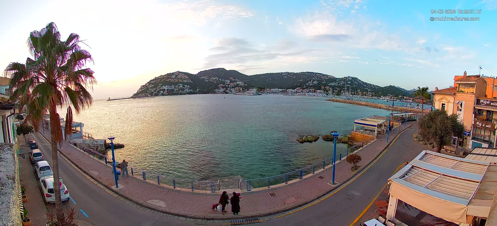 Two people walk a dog along a waterfront road at sunset, with a calm bay and distant hills visible in the background.
