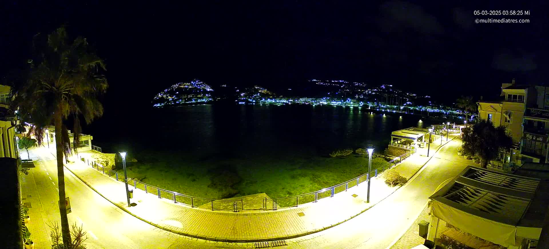 A nighttime, high-angle view shows a brightly lit waterfront promenade curving around a calm bay, with a hilltop town glowing with lights across the water.