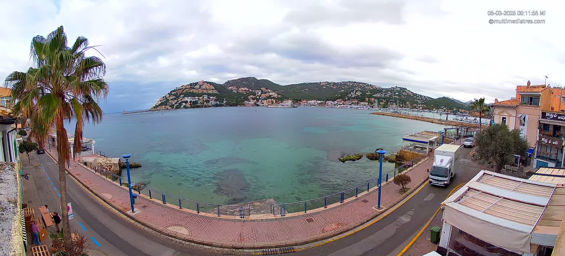 A seaside town with a calm, teal-colored bay, under a cloudy sky, is seen from an elevated vantage point.