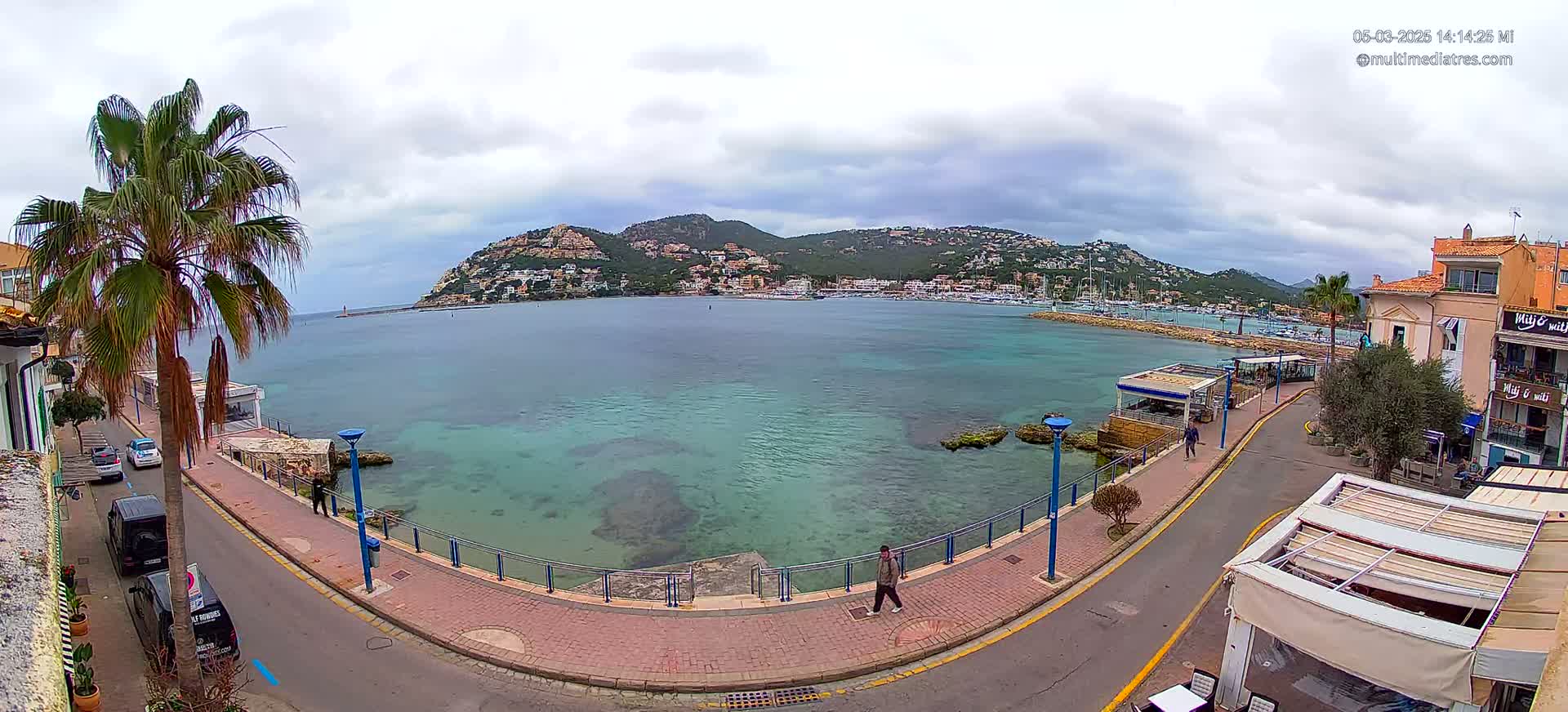 A coastal town scene on an overcast day shows a calm, teal-colored bay with a hill dotted with buildings in the background, bordered by a paved walkway and a street with parked cars.
