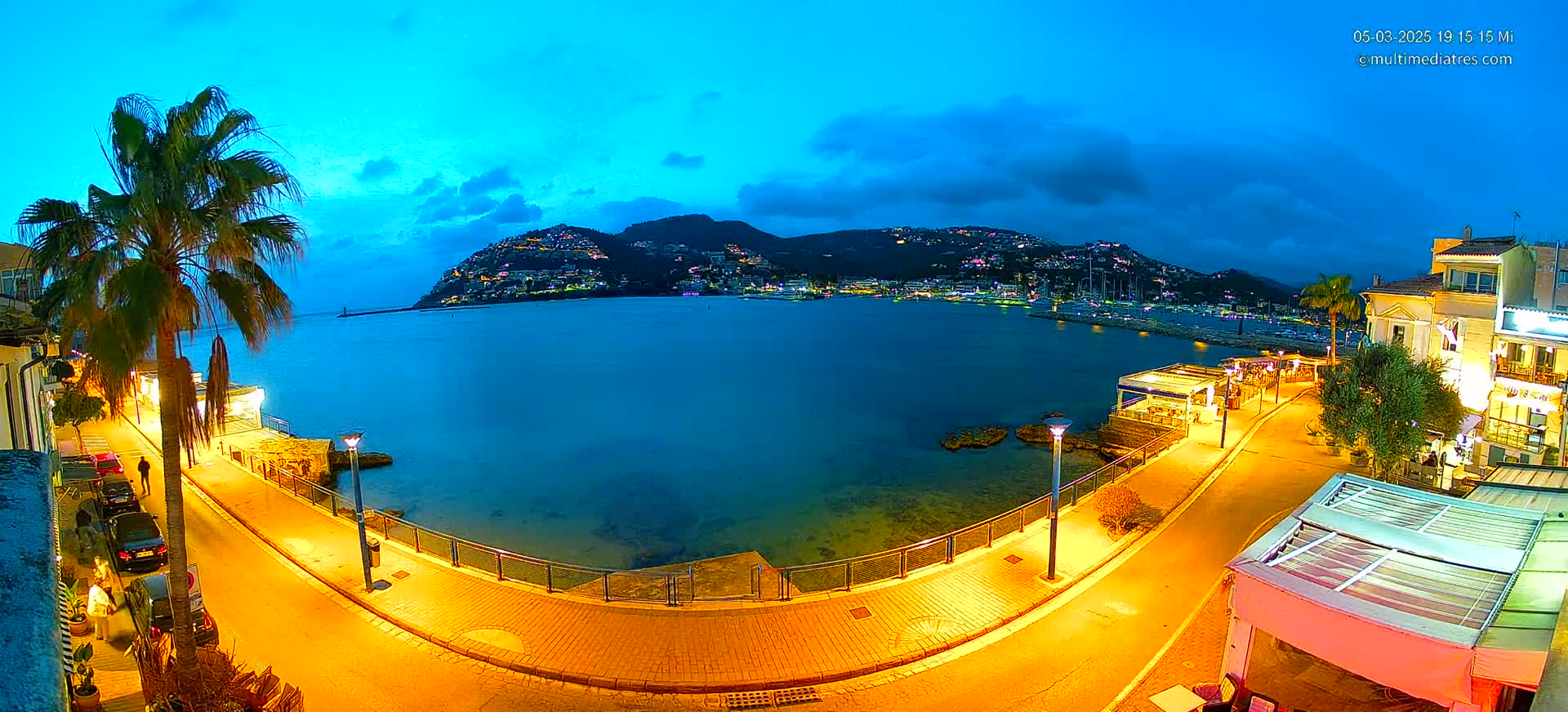 A brightly lit waterfront promenade at dusk overlooks a calm bay with a hilltop town in the background under a partly cloudy sky.