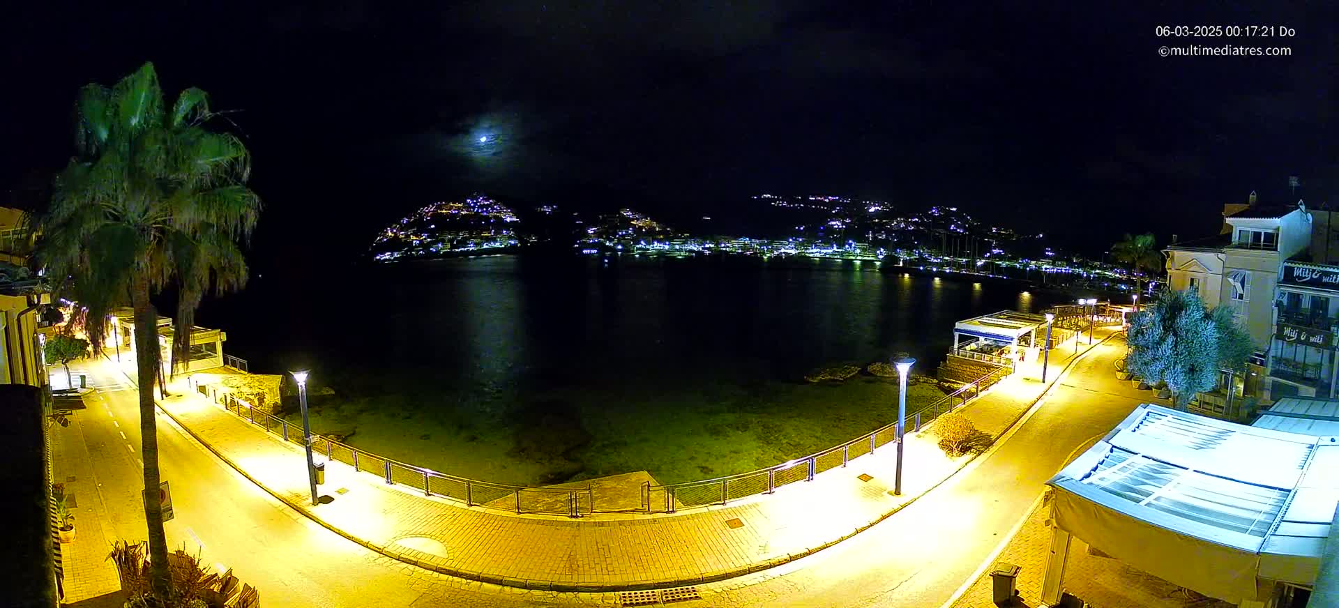A nighttime, moonlit view of a waterfront promenade illuminated by streetlights, with a calm body of water reflecting the lights of a town across the bay.