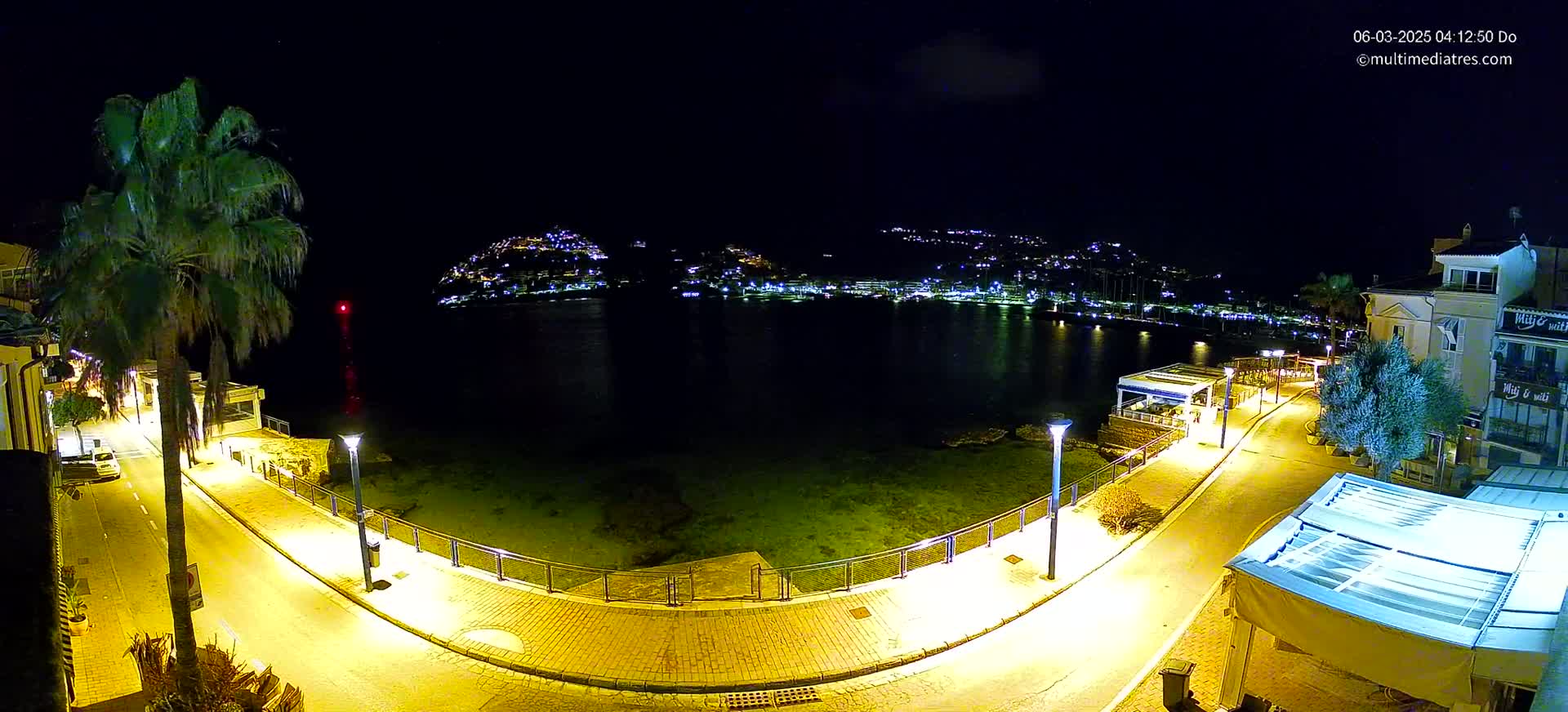 A nighttime, brightly lit seaside promenade curves around a calm bay with a town illuminated on the opposite shore.