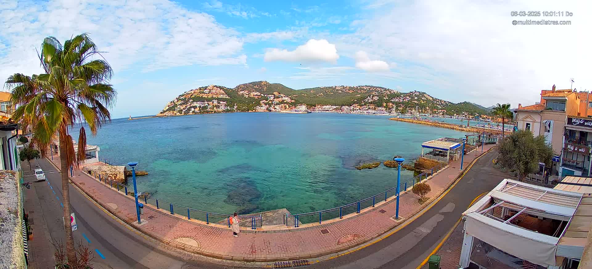 A partly cloudy day reveals a picturesque coastal town with a calm, clear turquoise bay, bordered by a paved walkway and buildings, and hills in the background.