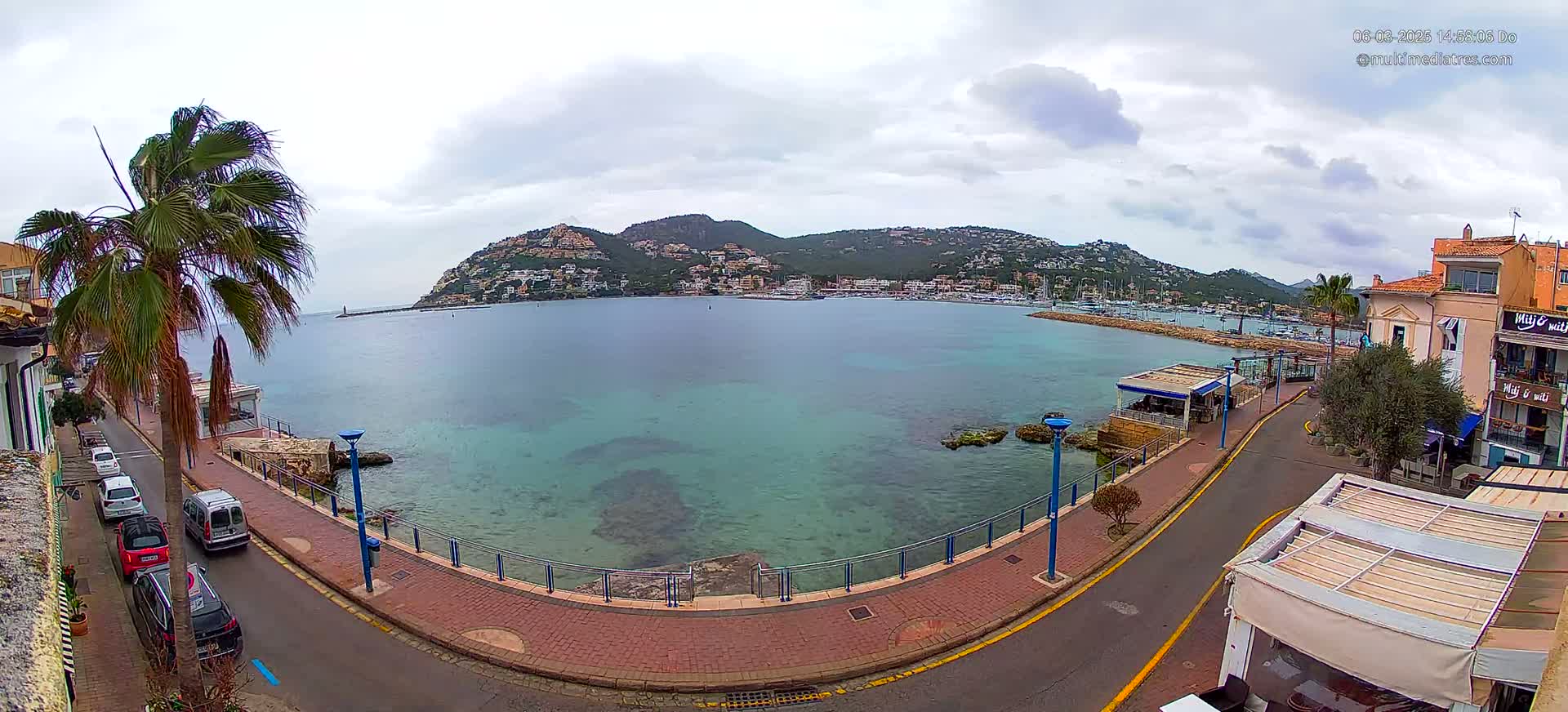 A seaside street with parked cars overlooks a calm, clear bay and a hill dotted with buildings under an overcast sky.