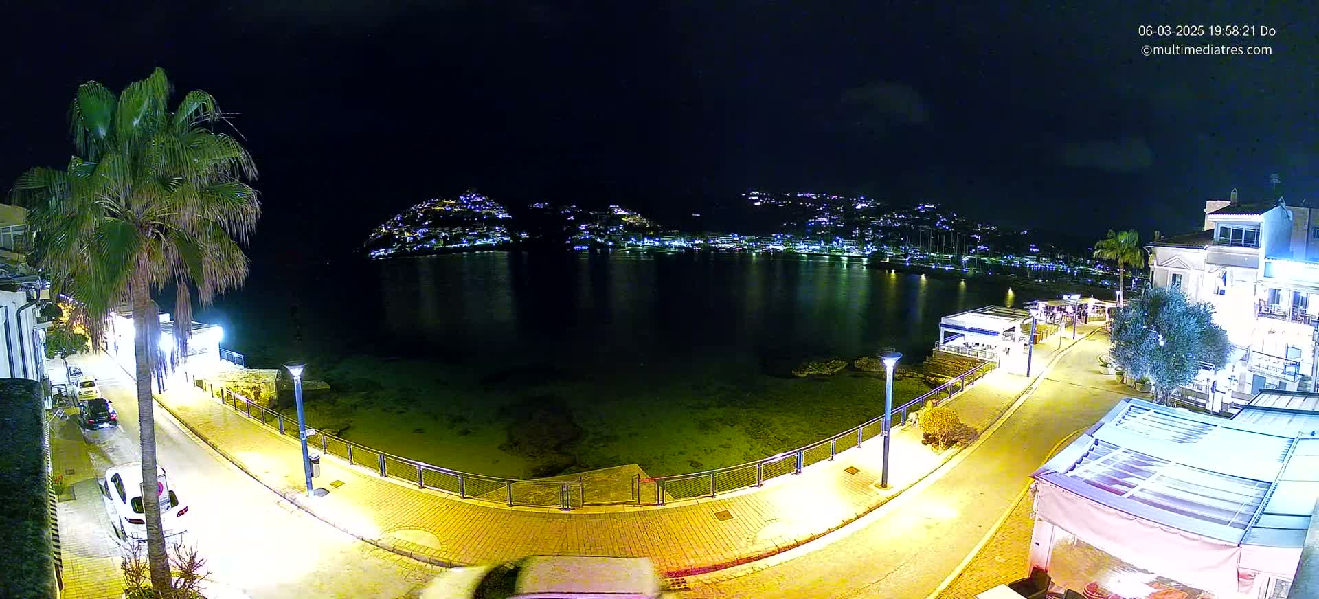 A nighttime view of a calm bay, with a town illuminated on the opposite shore, a palm tree and brightly lit waterfront promenade in the foreground.