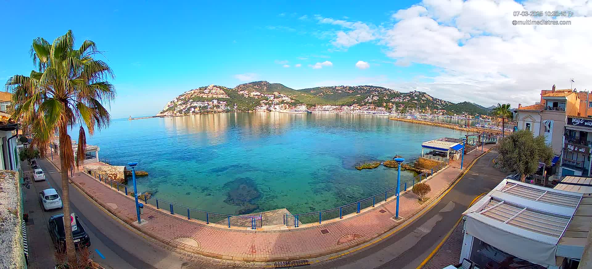 A clear, sunny day reveals a calm bay with clear turquoise water, a town nestled against a hillside, and a palm tree-lined street with cars parked along the waterfront.