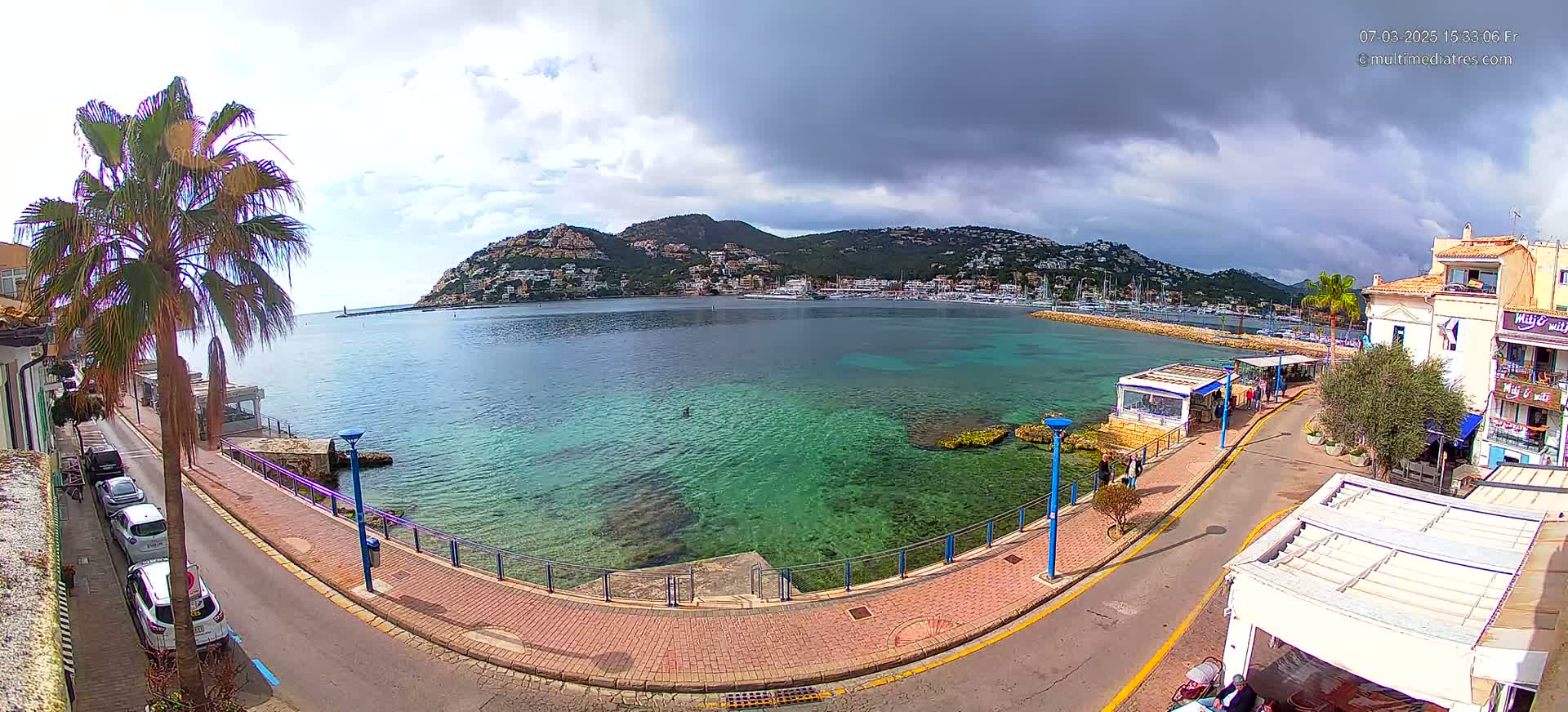 A partly cloudy day overlooks a clear, shallow bay with a town and hill in the background, viewed from a high vantage point above a paved walkway and street.