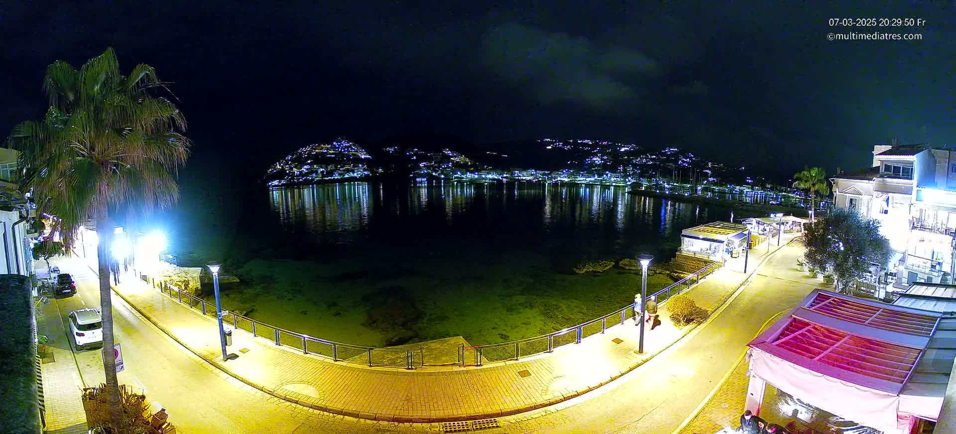 A nighttime view of a calm, illuminated harbor, bordered by a paved walkway, buildings, and palm trees.