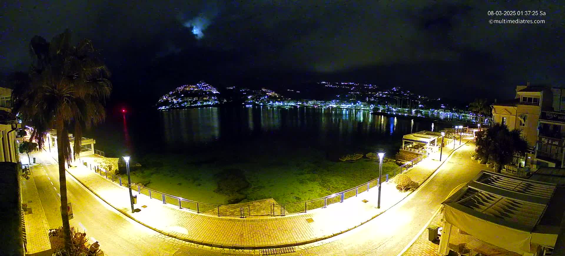 A nighttime, moonlit view of a waterfront promenade, illuminated by streetlights, overlooks a calm bay with a town's lights reflecting on the water.