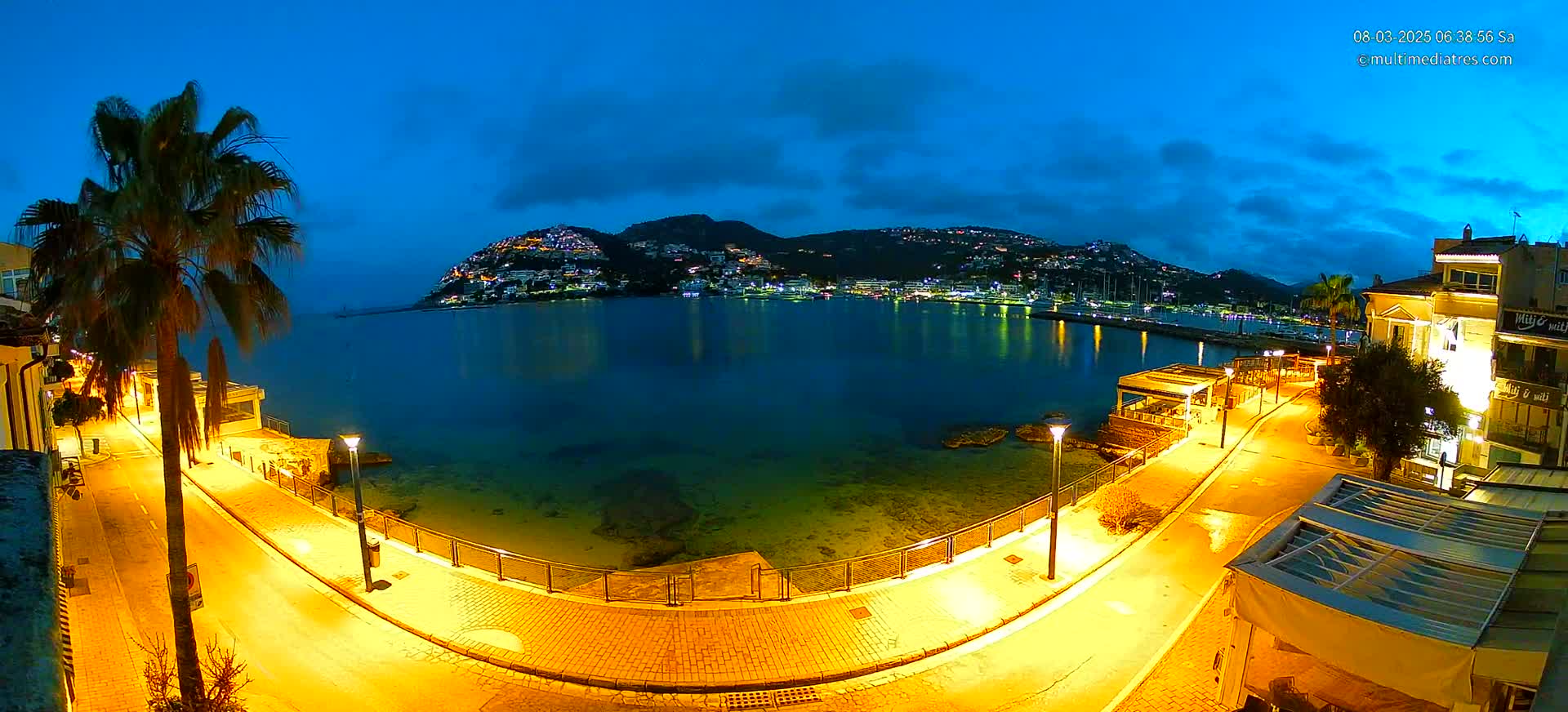 A nighttime coastal scene shows a calm bay reflecting the lights of a town nestled on a hillside, illuminated by streetlights along a paved walkway curving toward the water.