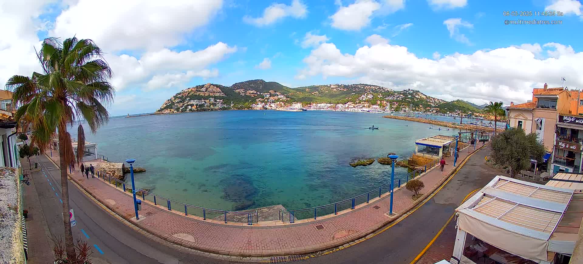 A seaside town with clear, turquoise water, a paved walkway, palm trees, and buildings on a hillside under a partly cloudy sky.