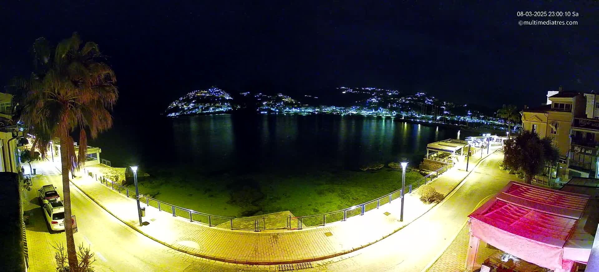 A nighttime, illuminated seaside promenade curves alongside a calm bay, overlooking a town's lights on a hill across the water.