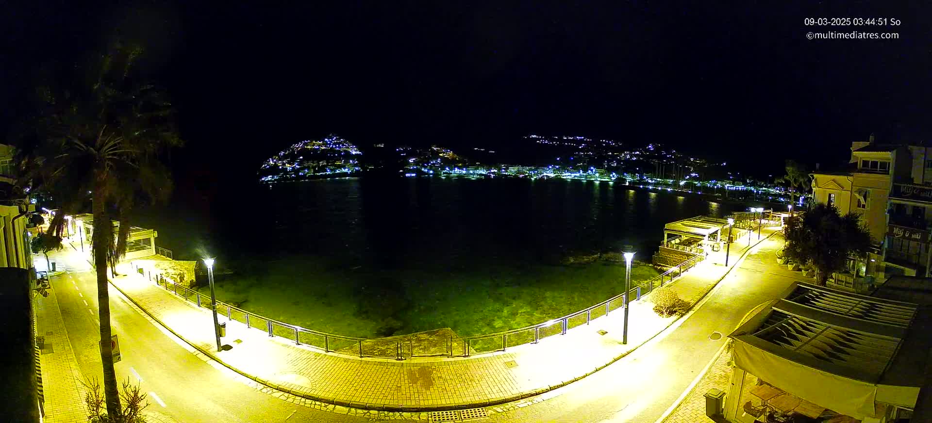 A nighttime, overhead view of a waterfront promenade illuminated by streetlights, overlooking a calm bay with a town lit up on the opposite shore.