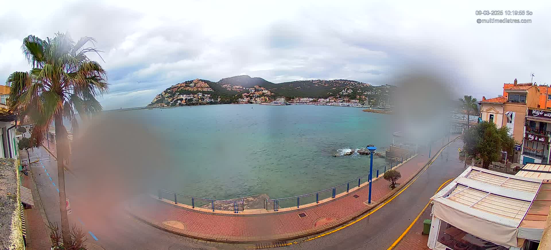 A seaside town on a cloudy day with a calm bay and hills covered in buildings in the background.