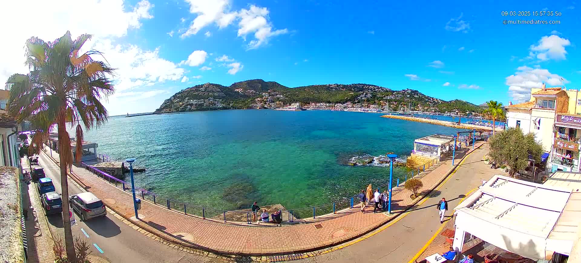 A clear, sunny day reveals a picturesque harbor town with turquoise water, a palm tree-lined promenade, parked cars, and a few people strolling along a paved walkway.