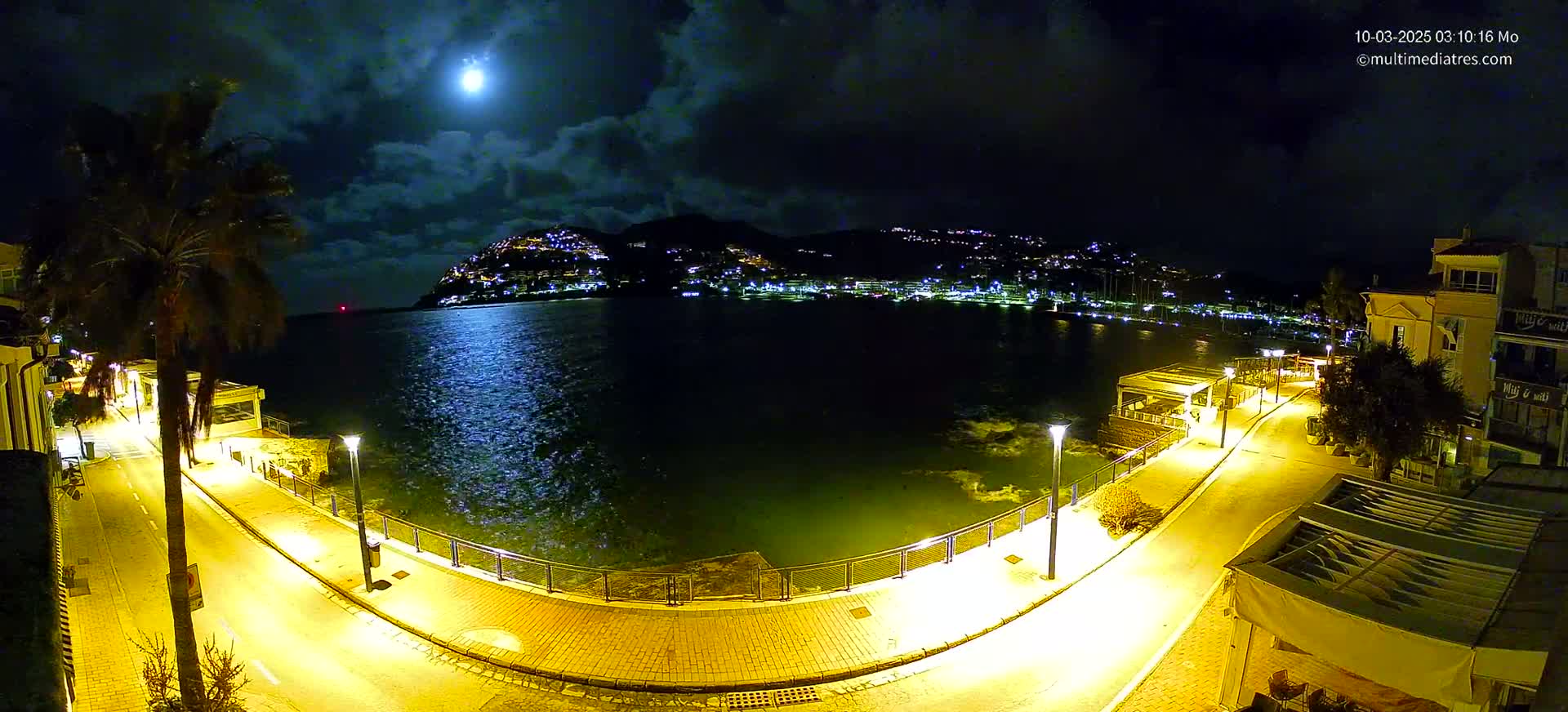 A moonlit night scene shows a brightly lit waterfront promenade curving around a dark body of water, with a hilltop town glowing in the distance under a partly cloudy sky.