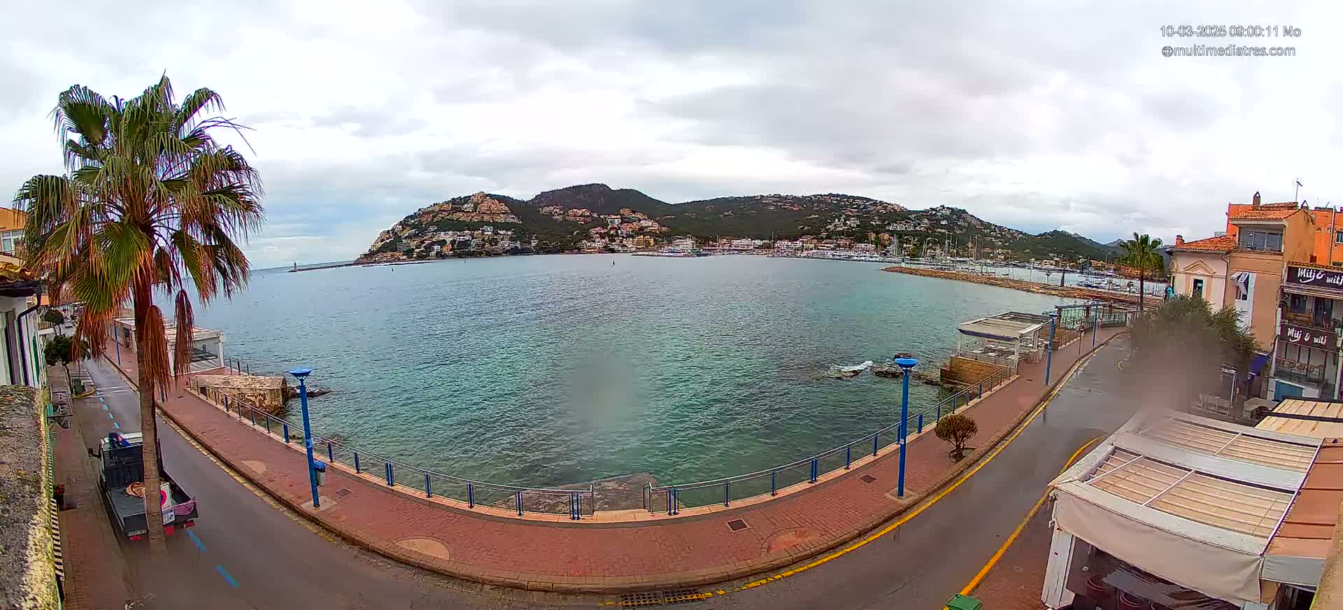 A seaside town, under an overcast sky, features a calm bay with a marina and hillside buildings in the background, seen from a street-level perspective.