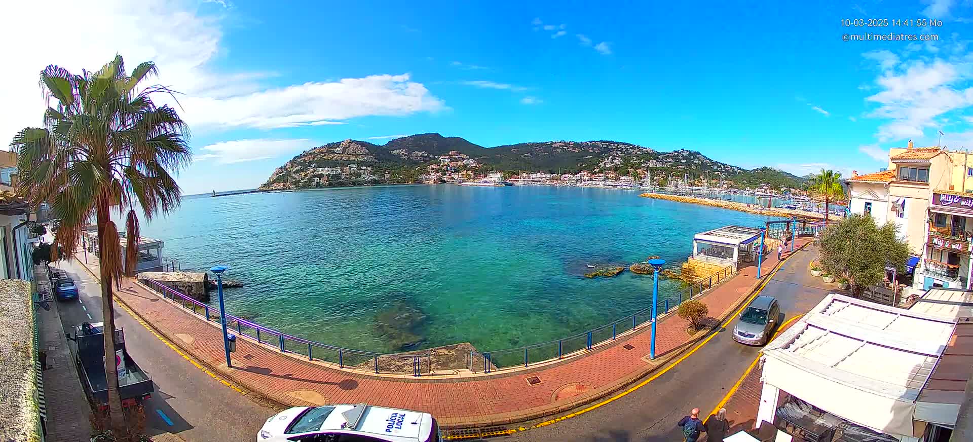 A clear, sunny day overlooks a calm, teal harbor with a hill covered in buildings in the background, viewed from a waterfront road lined with palm trees and buildings.