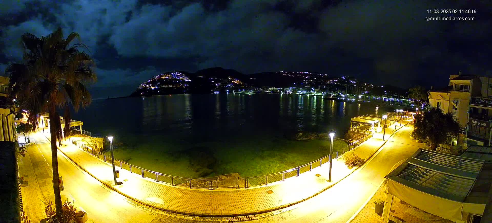 A nighttime coastal scene shows a town illuminated on a hillside across a calm, dark bay, with a brightly lit paved walkway curving along the water's edge, under a partly cloudy sky.