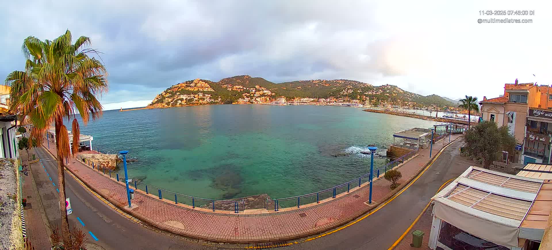 A coastal town with a calm, teal-colored bay, overlooked by a hillside of buildings under a partly cloudy sky.
