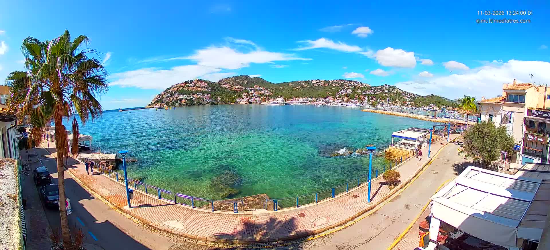 A sunny day shows a waterfront walkway beside clear teal water, with a hill covered in buildings in the background.