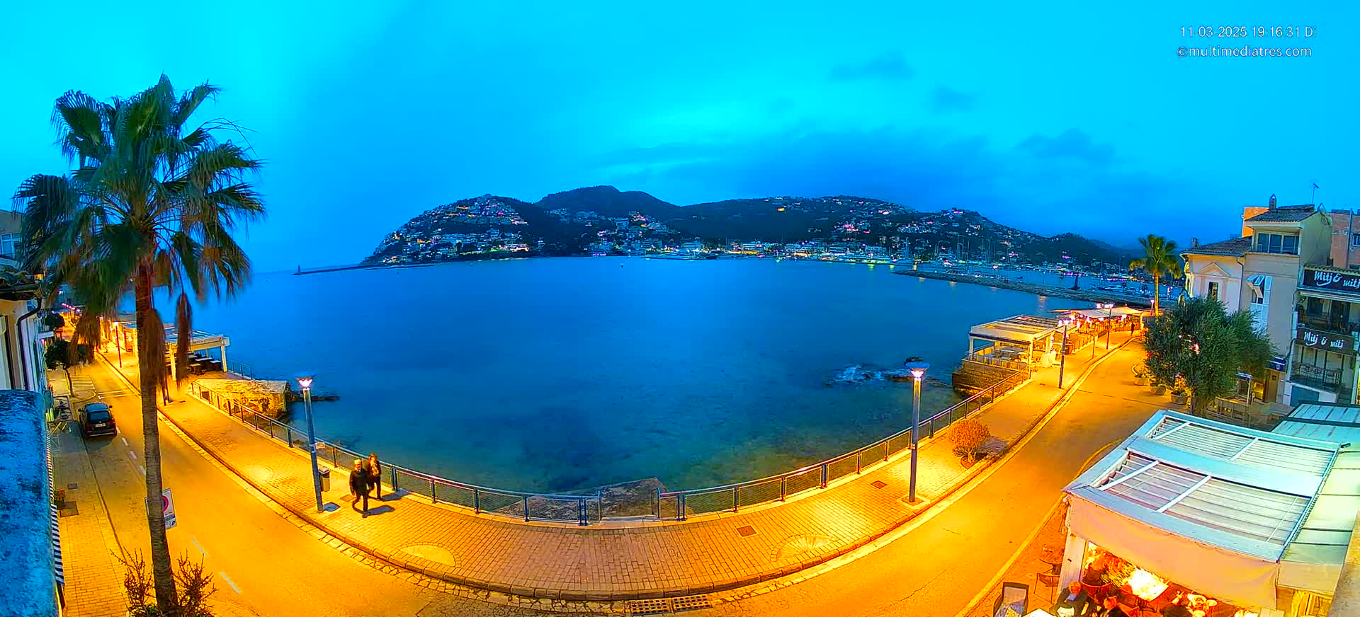 A seaside town at dusk, illuminated by streetlights, overlooks a calm body of water with a hill covered in lights in the background.