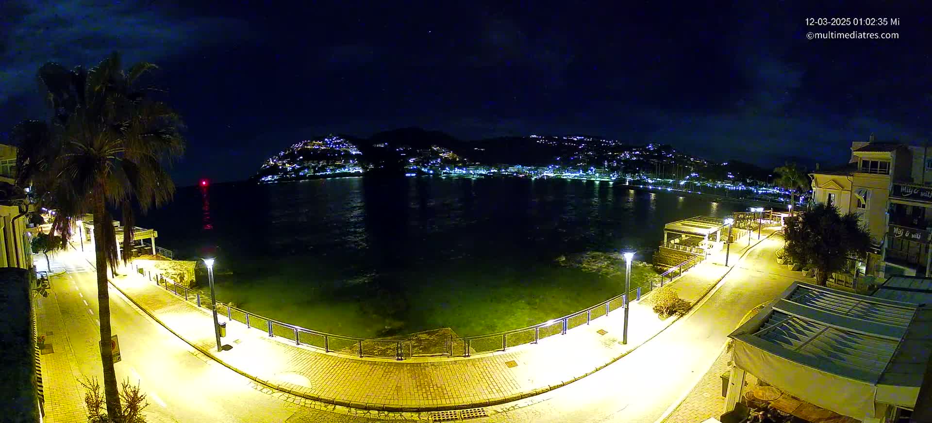 A nighttime view of a calm bay with a hilltop town illuminated across the water, seen from a waterfront promenade with palm trees and streetlights.