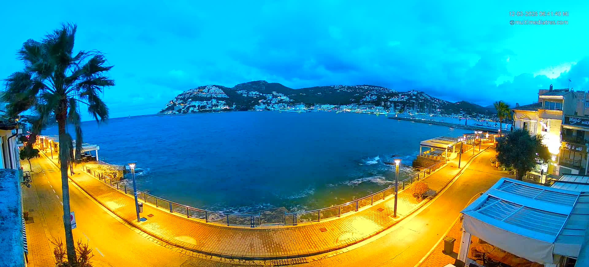 A seaside promenade at dusk, illuminated by streetlights, curves around a calm bay with a hilltop town visible in the background under a mostly clear, twilight sky.