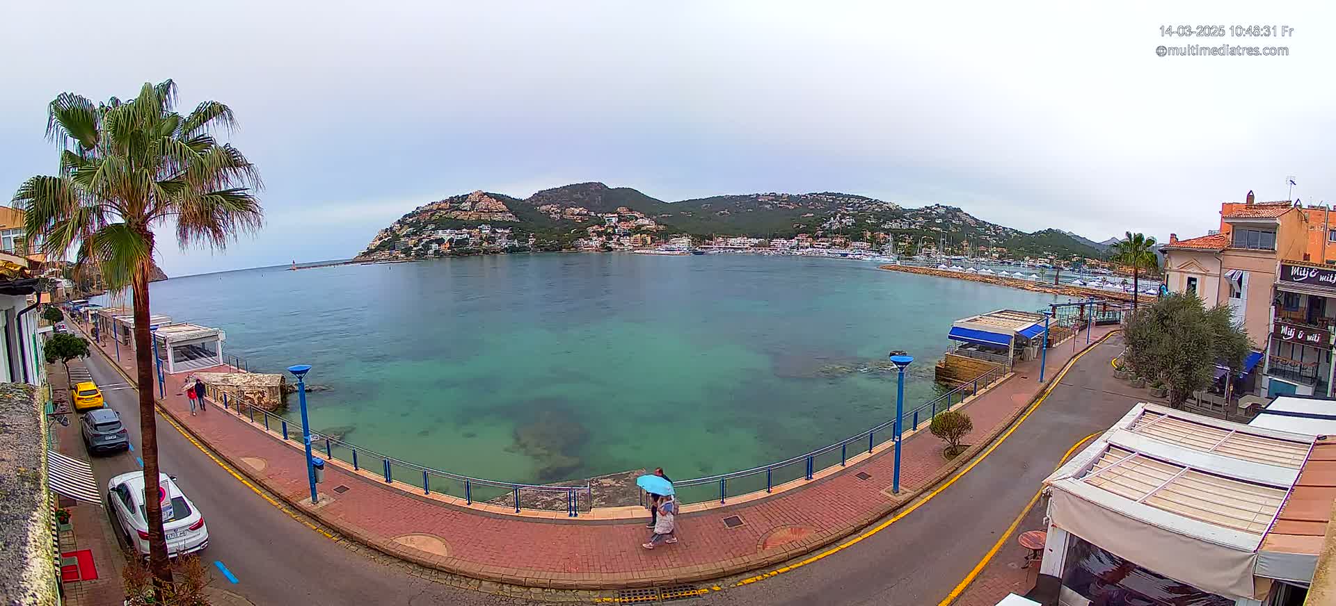A coastal town with a calm, clear bay, buildings lining the shore, and a couple walking along a paved walkway under an overcast sky.