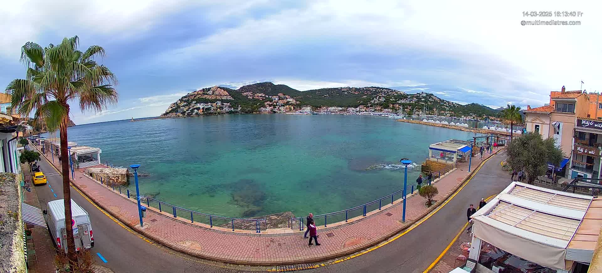 A waterfront scene with clear, teal water, a hill covered in buildings in the background, and a partly cloudy sky.