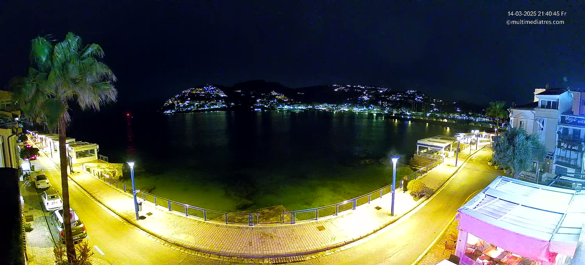 A nighttime view of a waterfront promenade, illuminated by streetlights, with a calm body of water reflecting the lights of buildings on a hill in the background.
