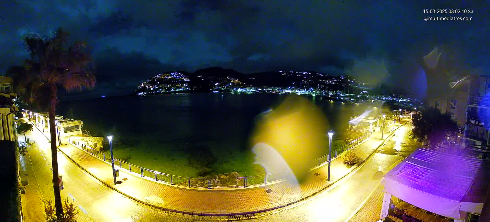 A rain-slicked, nighttime waterfront path curves toward a calm bay under a dark, cloudy sky, illuminated by streetlights and the distant lights of a town nestled on a hillside.